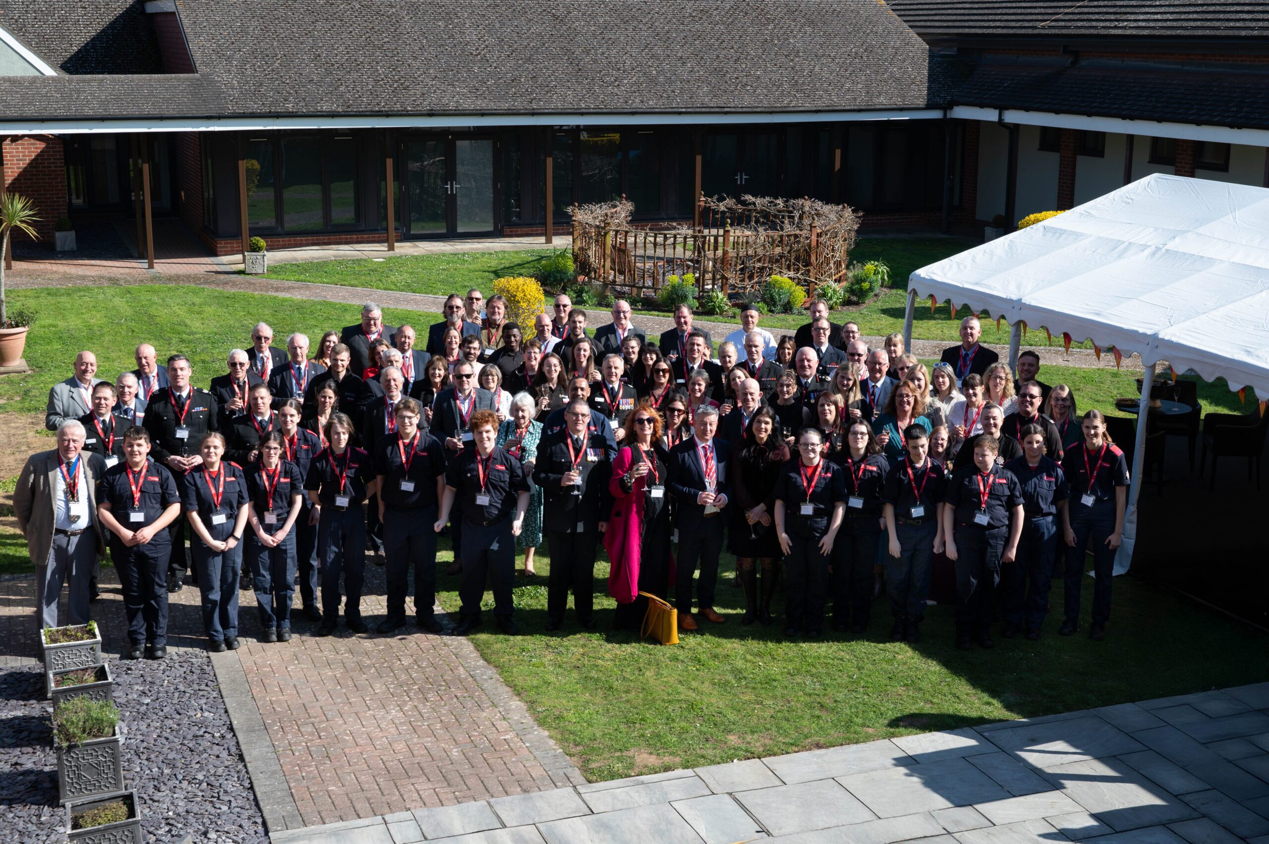 A large group of people wearing black outfits and red lanyards pose together outdoors in a courtyard, with a white event tent on the right and buildings in the background under bright sunlight.