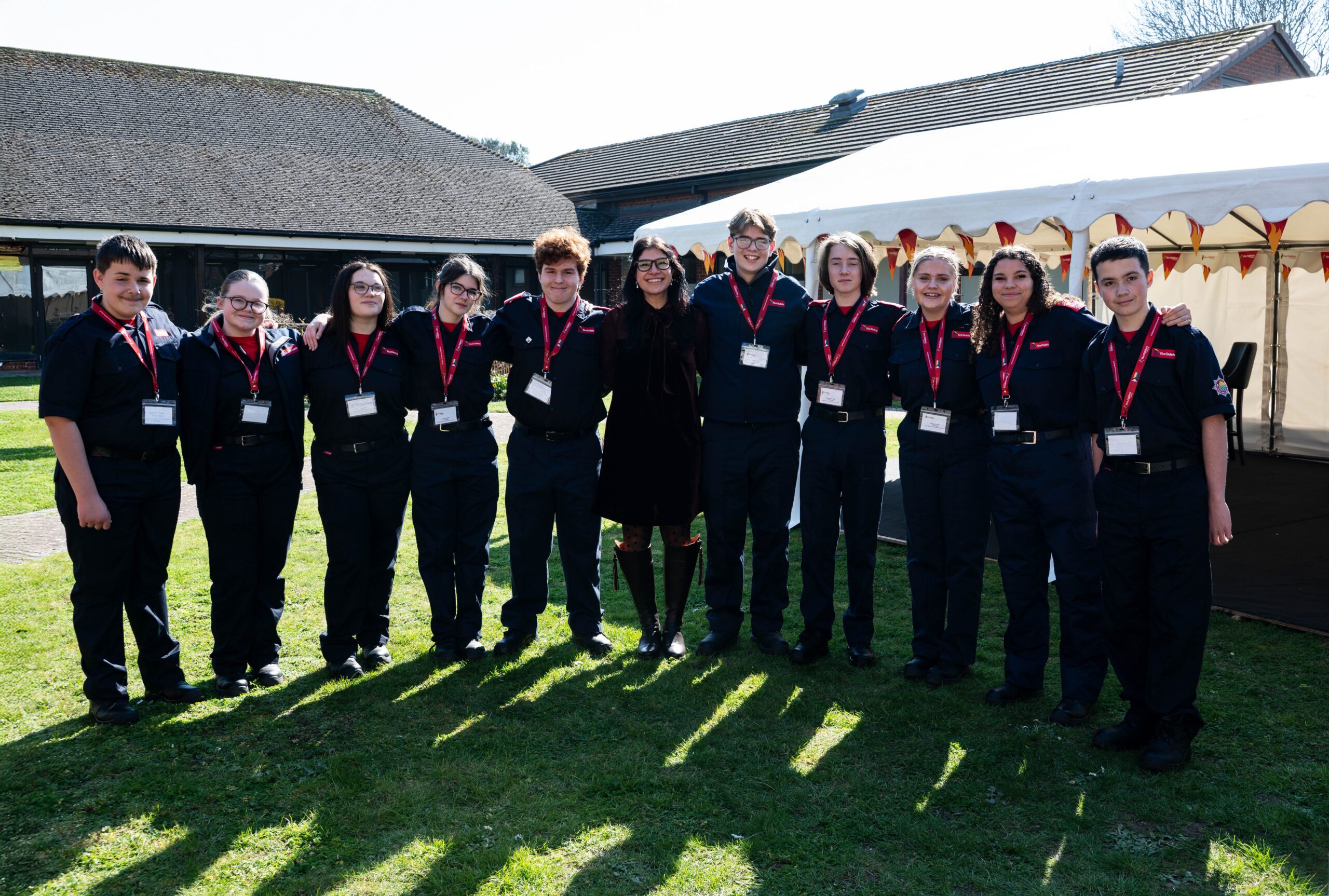A group of twelve people wearing matching black uniforms and red lanyards stand in a row outdoors on grass, smiling for the camera. A white tent and brick building are in the background.