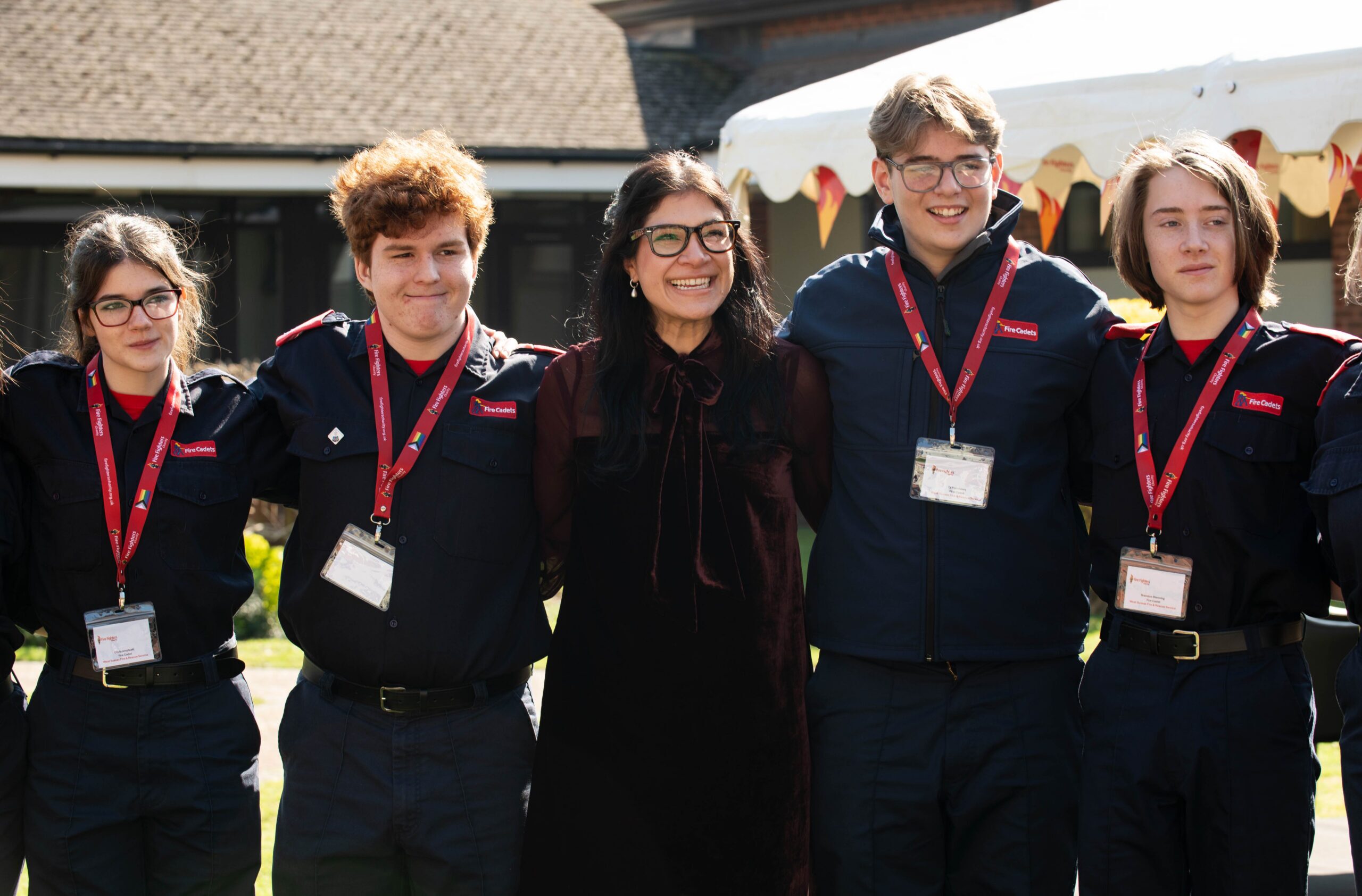 A group of young people in blue uniforms with red lanyards stand outside, smiling with a woman in black glasses and a dark dress, under bright sunlight near a tent.