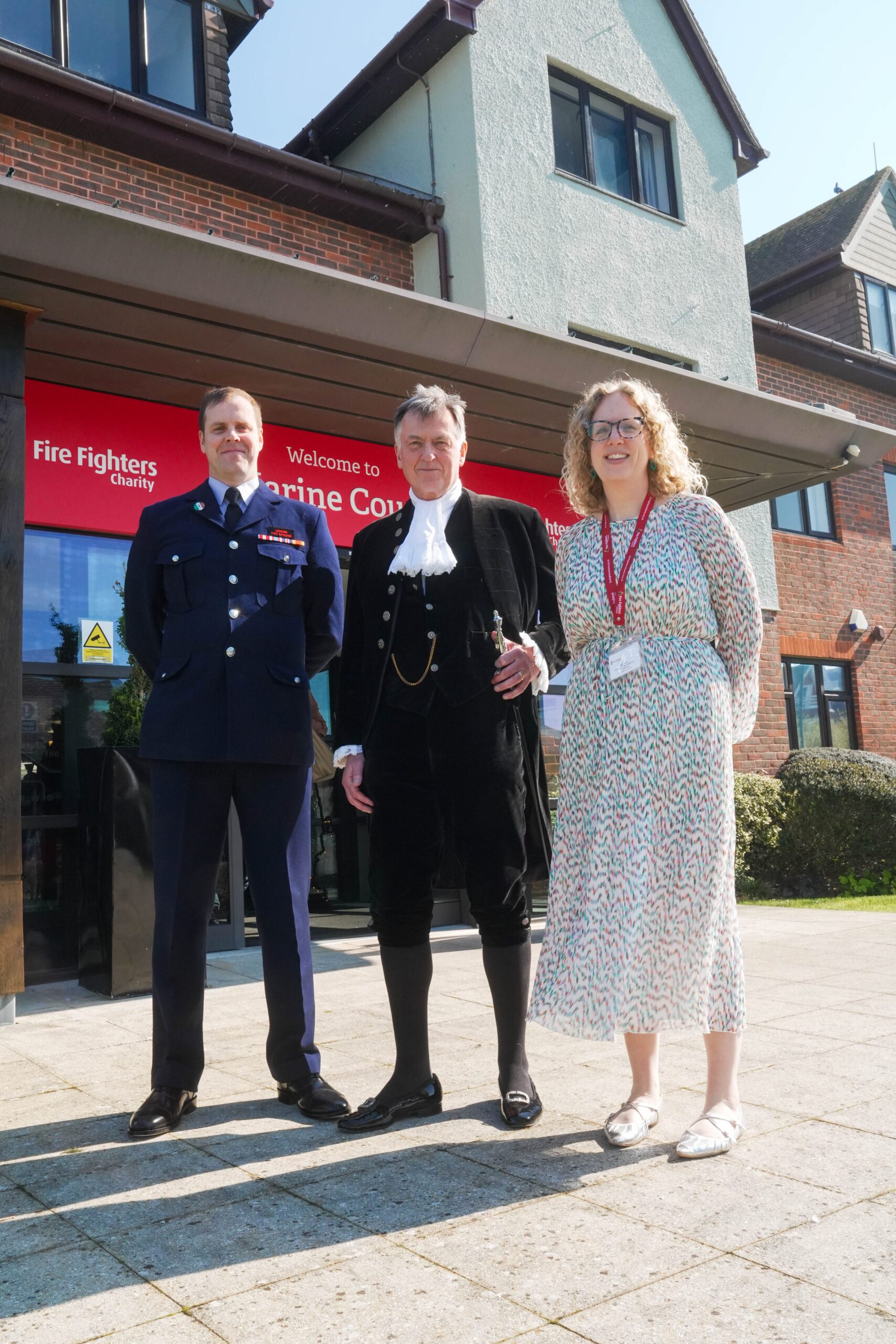 Three people stand outside a building with a sign reading "Fire Fighters Charity." One wears a formal uniform, one is dressed in traditional regalia, and one wears a patterned dress and lanyard.