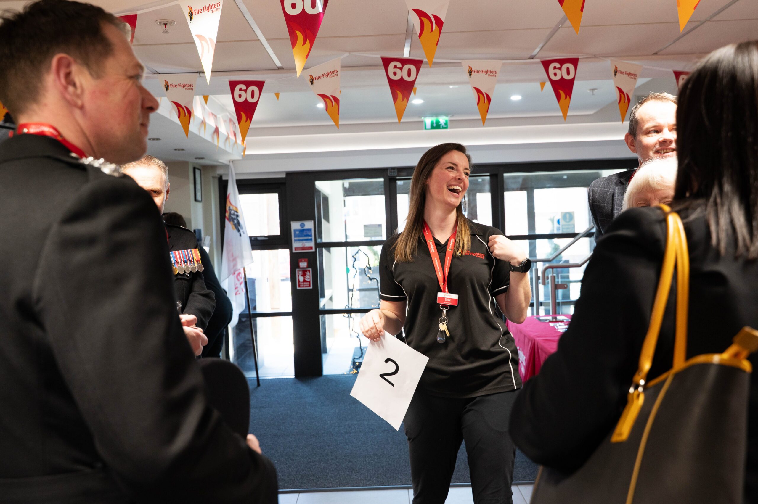 A woman holding a paper with the number 2 smiles and talks with a group of people at an indoor event decorated with "60" bunting.