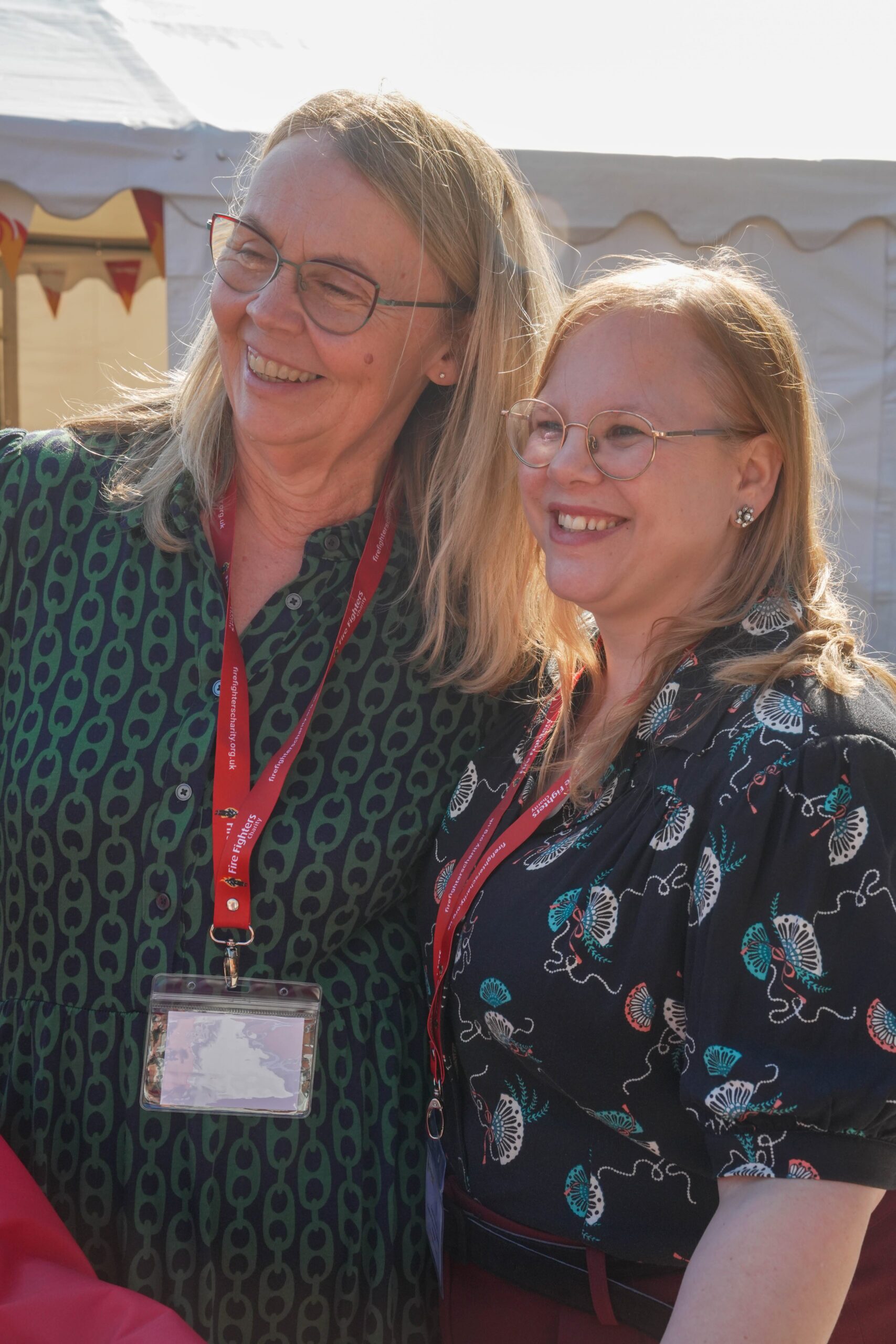 Two smiling women with glasses stand close together outdoors, both wearing patterned shirts and red lanyards with name badges, in front of a white event tent.