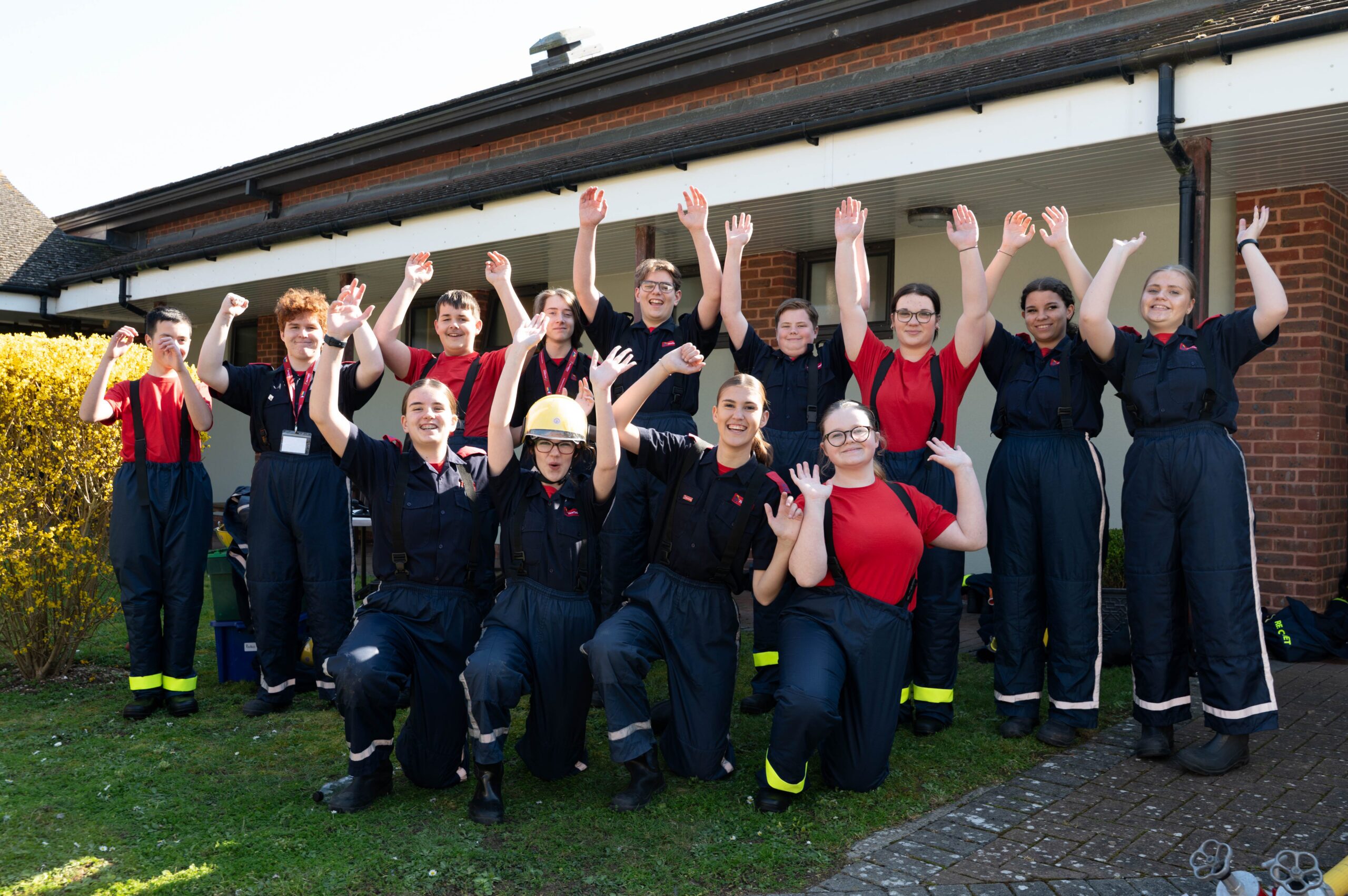 A group of firefighters in uniform kneel on grass outside a building, smiling and raising their arms in celebration. Some wear helmets, while others have red undershirts. Bright sunlight and a bush are visible.