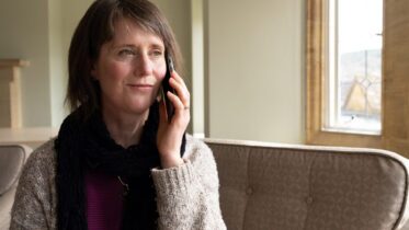 A woman with short brown hair sits on a sofa indoors, smiling gently while talking on a smartphone. She wears a cozy sweater, a dark scarf, and looks toward a window letting in natural light.