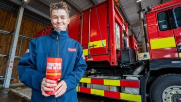 A young person in a blue jacket holds a red charity collection tub, smiling in front of a bright red fire truck inside a fire station.