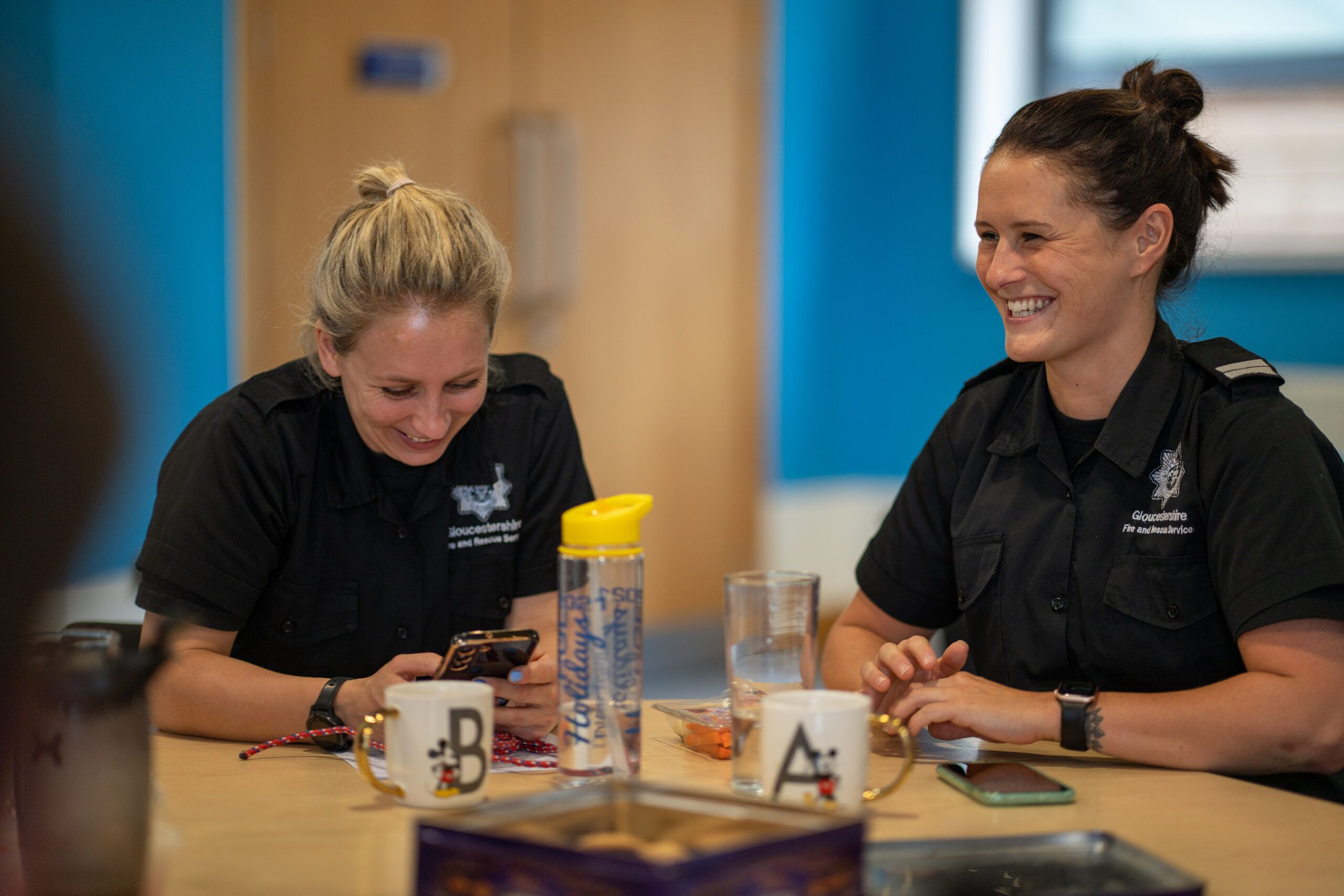 Two women in dark uniforms sit at a table, smiling and laughing. One is looking at her phone while the other looks at her. Mugs and a water bottle are on the table. The setting appears casual and relaxed.