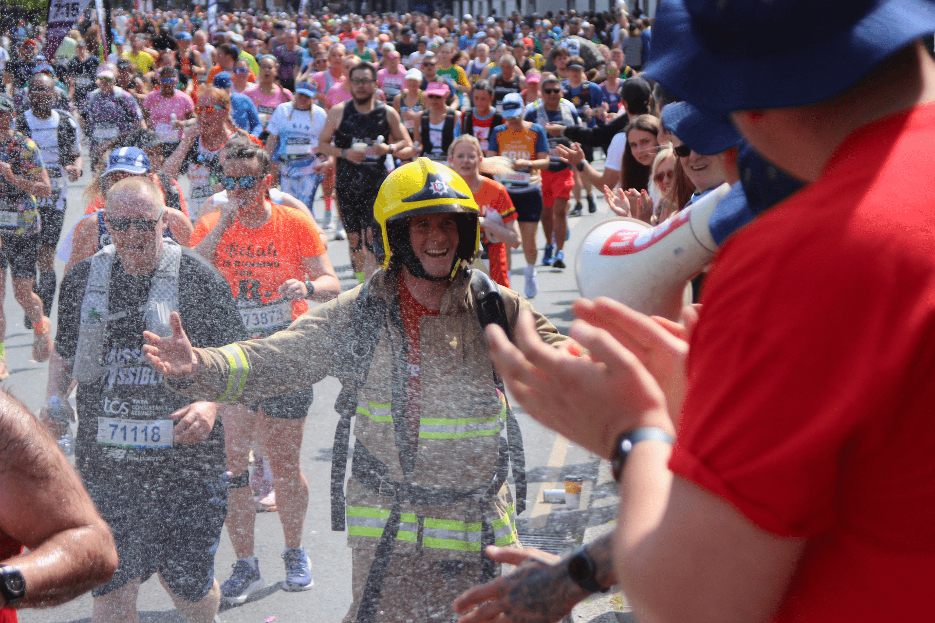 A firefighter in uniform and helmet runs in a crowded marathon, smiling as confetti or water is sprayed. Runners and spectators in colorful outfits cheer and clap along the street.