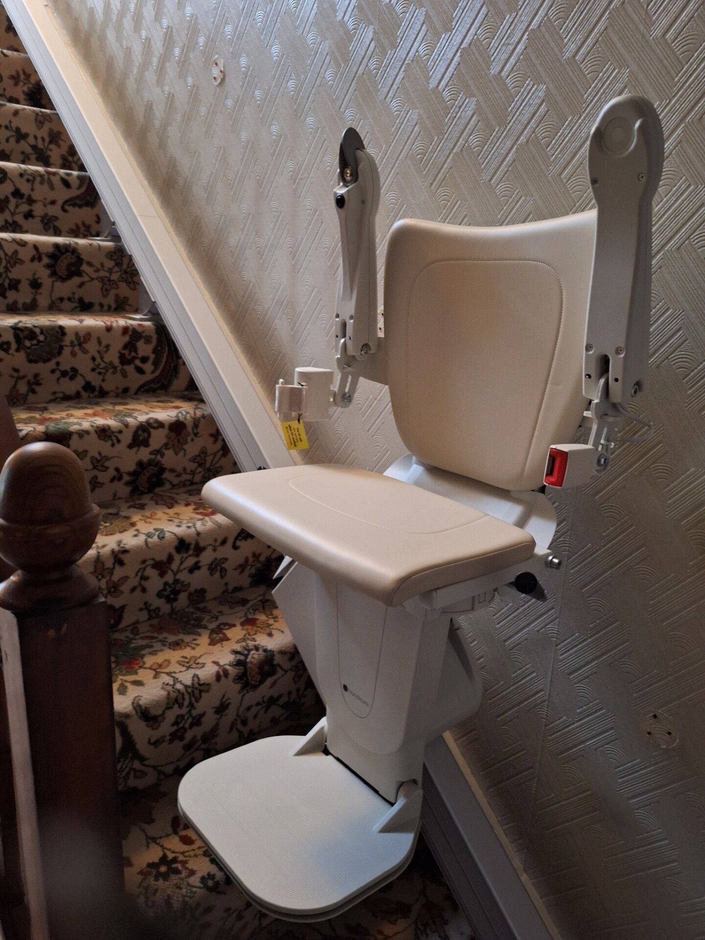 A cream-colored stairlift chair is installed along a carpeted staircase with a patterned banister and textured wallpaper on the wall beside it. The chair’s armrests are raised, and footrest is folded down.