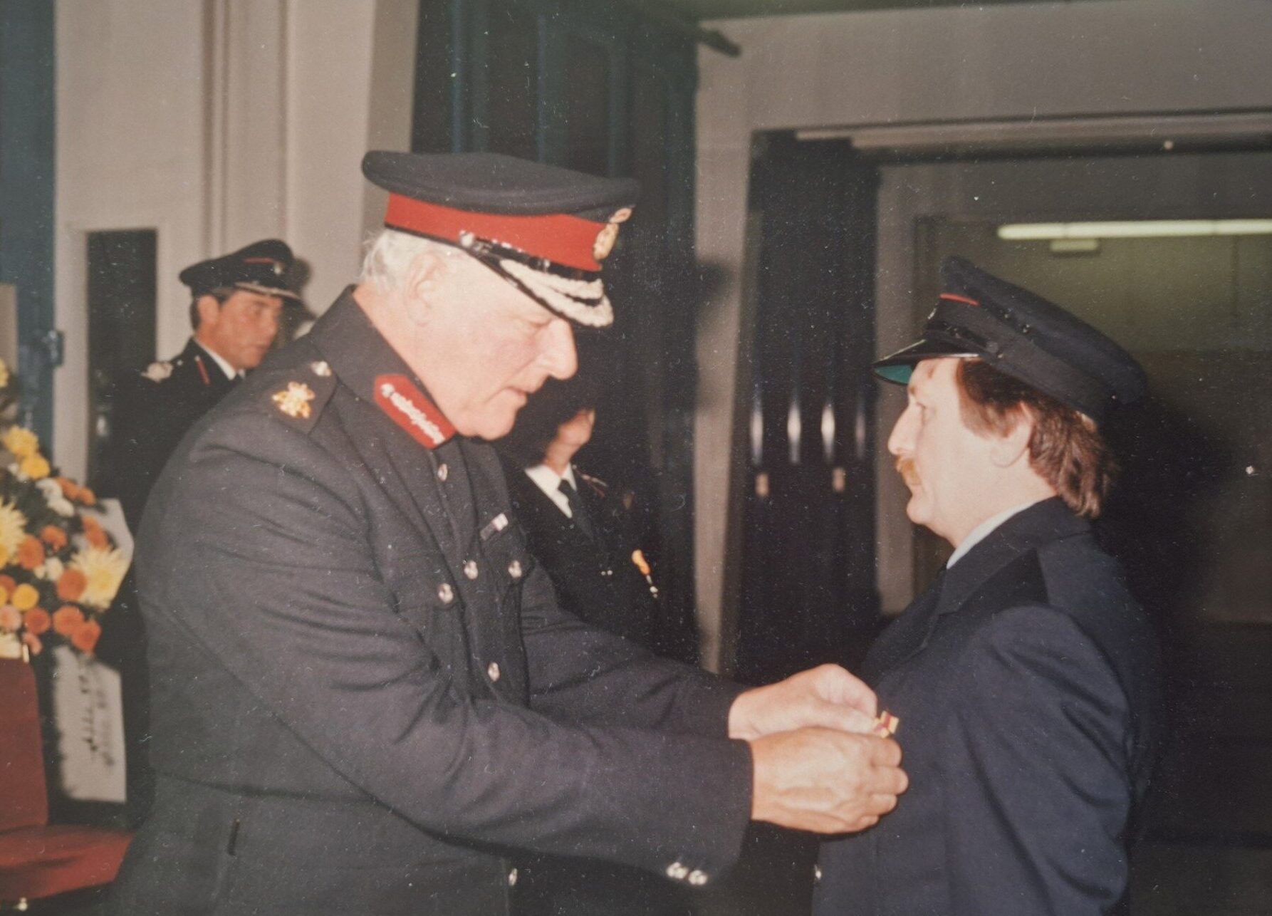 Two people in formal uniforms, including hats, shake hands indoors. One person has white hair and the other has brown hair. A bouquet of flowers can be seen in the background.