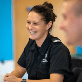 A smiling woman in a dark uniform sits at a table, holding a mug. The background is softly blurred, and another person is partially visible in the foreground.