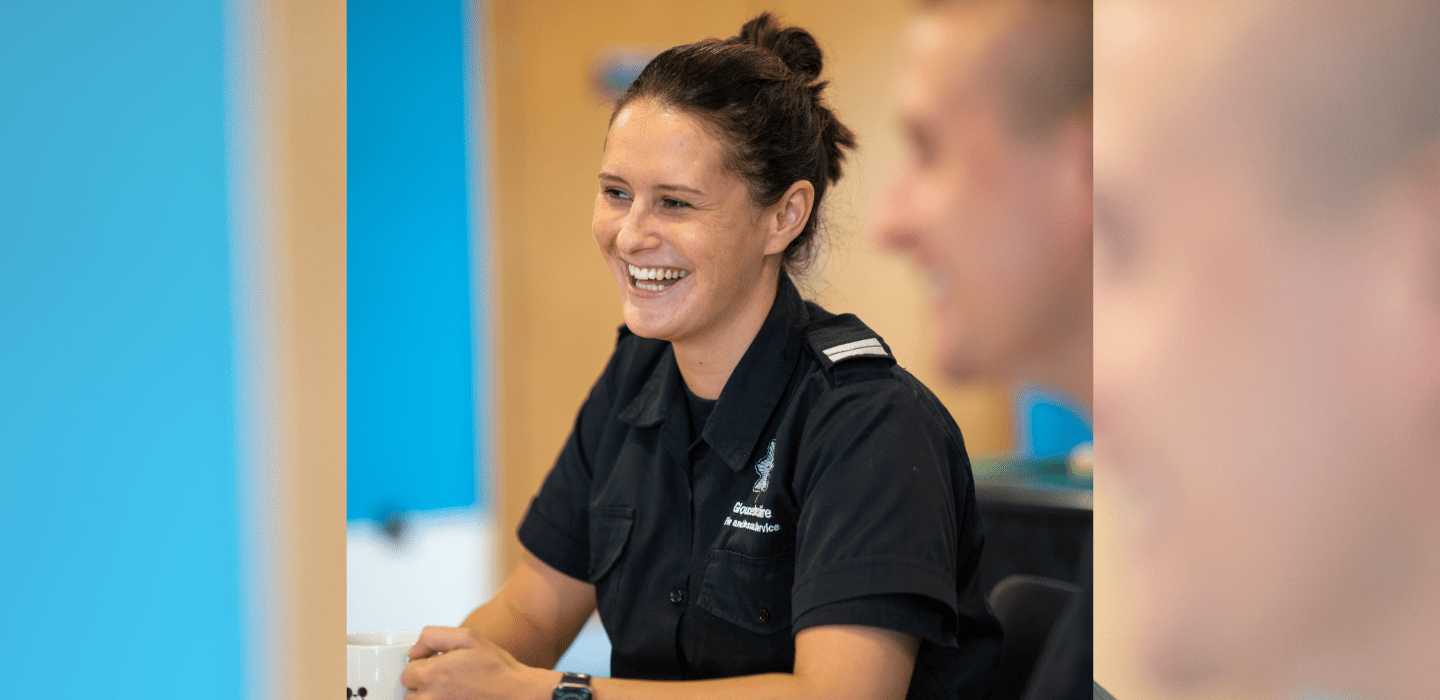 A smiling woman in a dark uniform sits at a table, holding a mug. The background is softly blurred, and another person is partially visible in the foreground.
