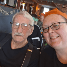An older man with glasses and a woman wearing glasses and a paper crown smile for a selfie in a festive, decorated room with a 