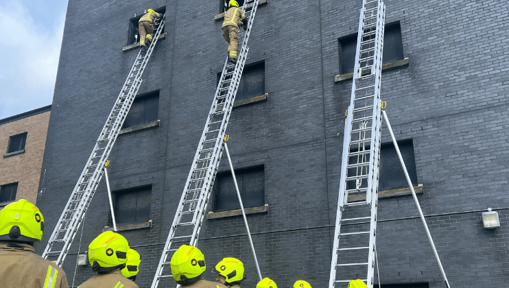 Firefighters wearing bright yellow helmets stand on the ground, watching as two firefighters climb tall ladders leaning against a grey brick building during a training exercise.