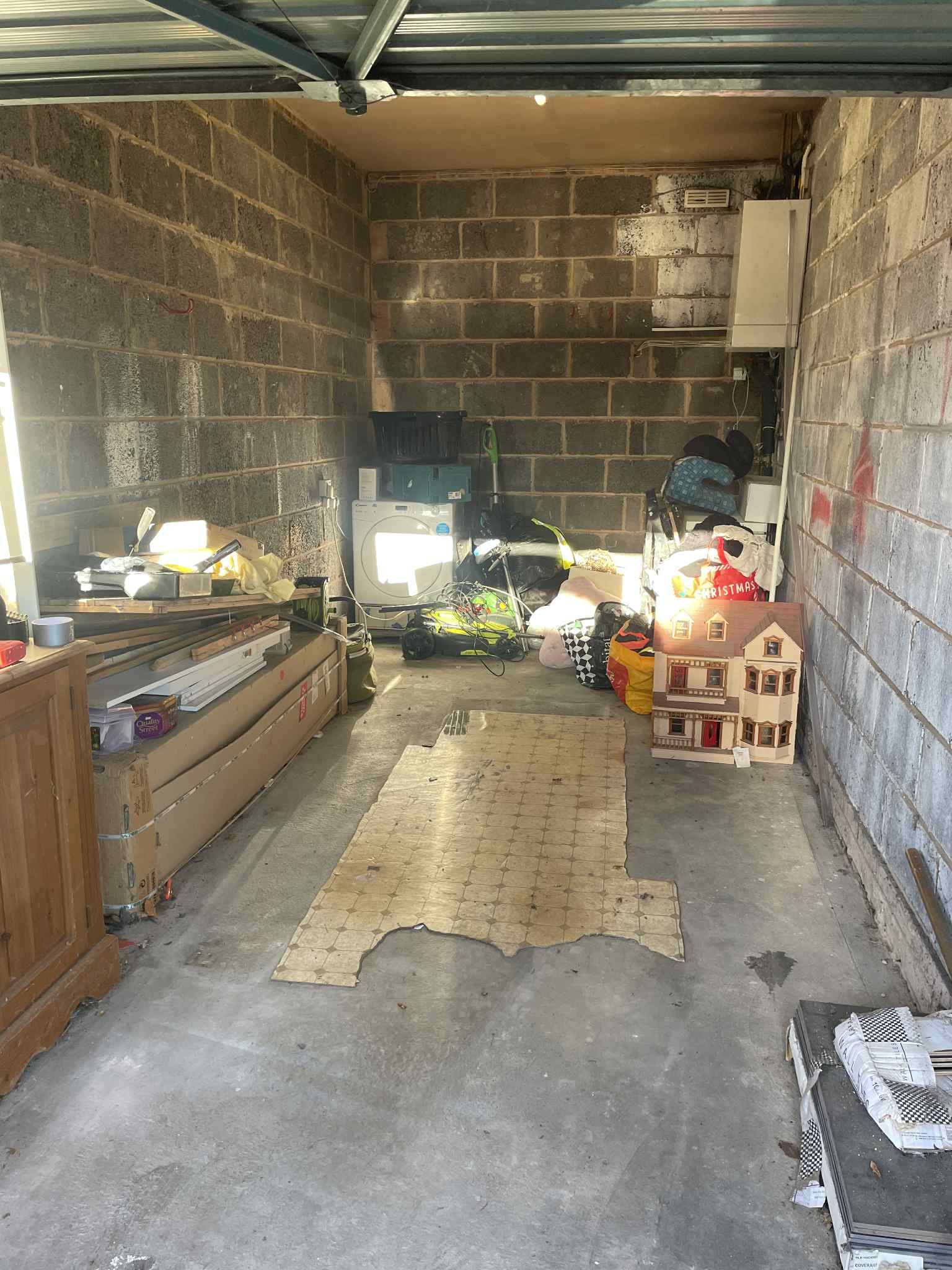 A cluttered garage with exposed brick walls, various boxes, tools, a toy dollhouse, and scattered household items; sunlight streams in, illuminating the back wall and some objects on the floor.