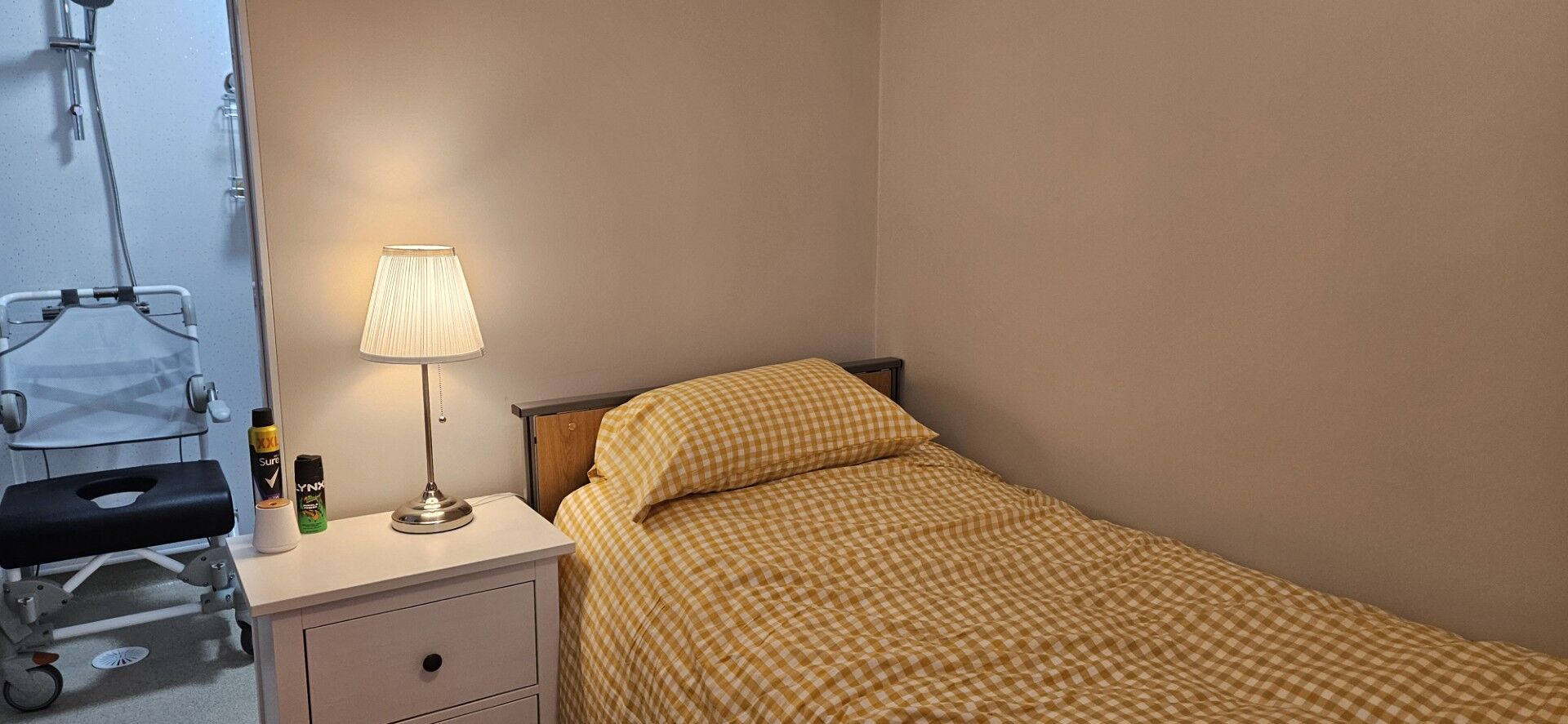 A small bedroom with a single bed covered in yellow checkered bedding, a white nightstand with a lamp and toiletries, and a glimpse of a bathroom with a shower chair visible through an open door.