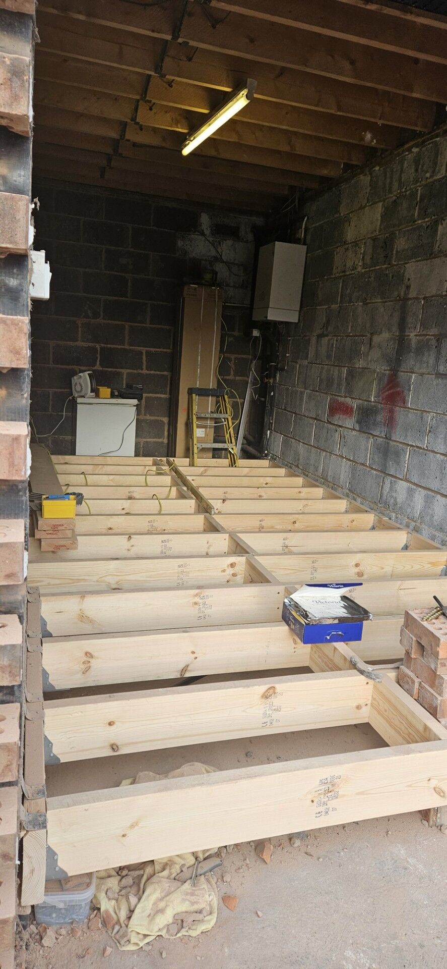A garage with exposed brick and block walls is being renovated. Wooden floor joists are installed, with tools and materials resting on them. The ceiling shows exposed beams, and a fluorescent light hangs overhead.