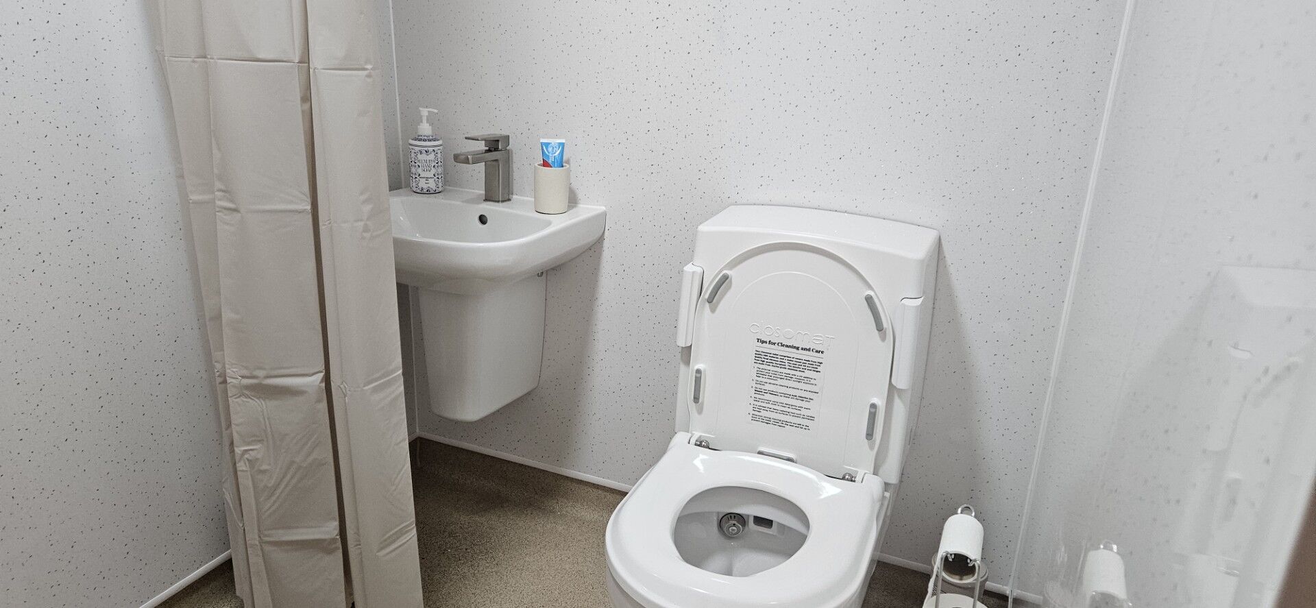 A clean, white bathroom with a toilet, a small sink, a shower curtain, soap dispenser, toilet paper, and a cup with toothbrushes on the sink. The overall space is simple and tidy.