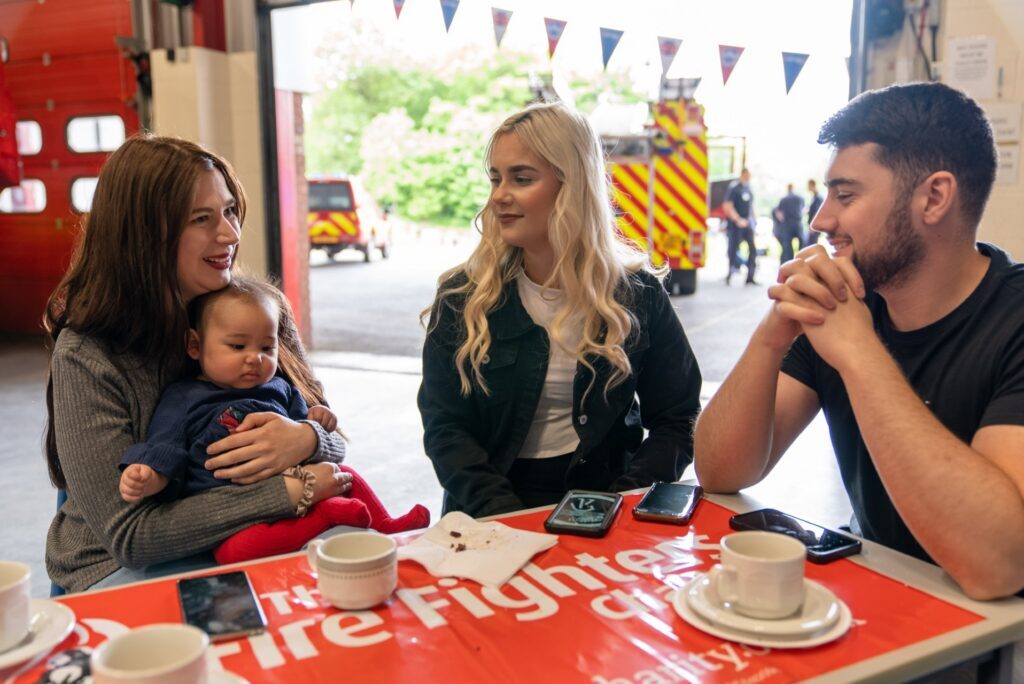 Three adults and a baby sit around a table with drinks and cellphones, smiling and talking. Fire engines are visible outside, and a red "Fire Fighters Charity" banner covers the table.