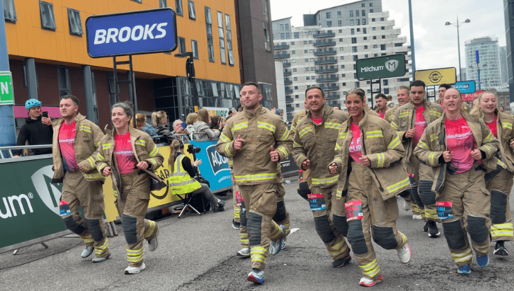 A group of people in firefighter uniforms run together during a race in a city, smiling and cheered on by spectators behind barriers and banners. Modern buildings and event signage are visible in the background.