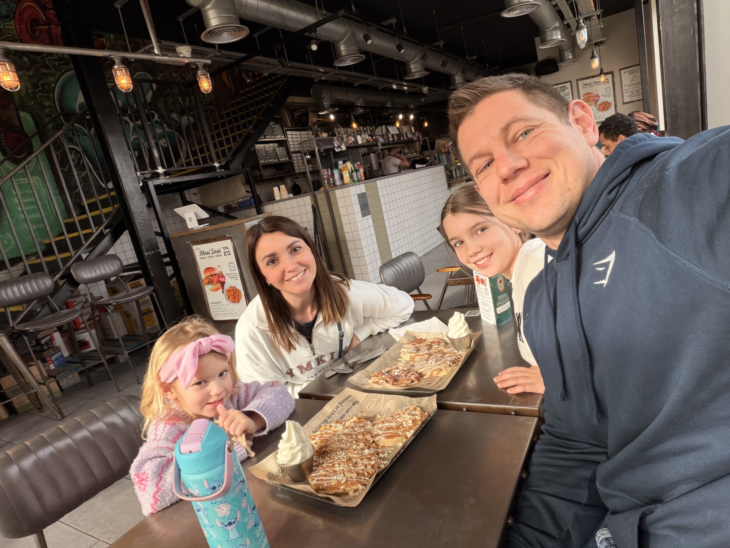 A family of four sits at a table in a café, smiling at the camera. In front of them are plates with waffles topped with sauce and whipped cream. The café has industrial décor with stools and a staircase in the background.