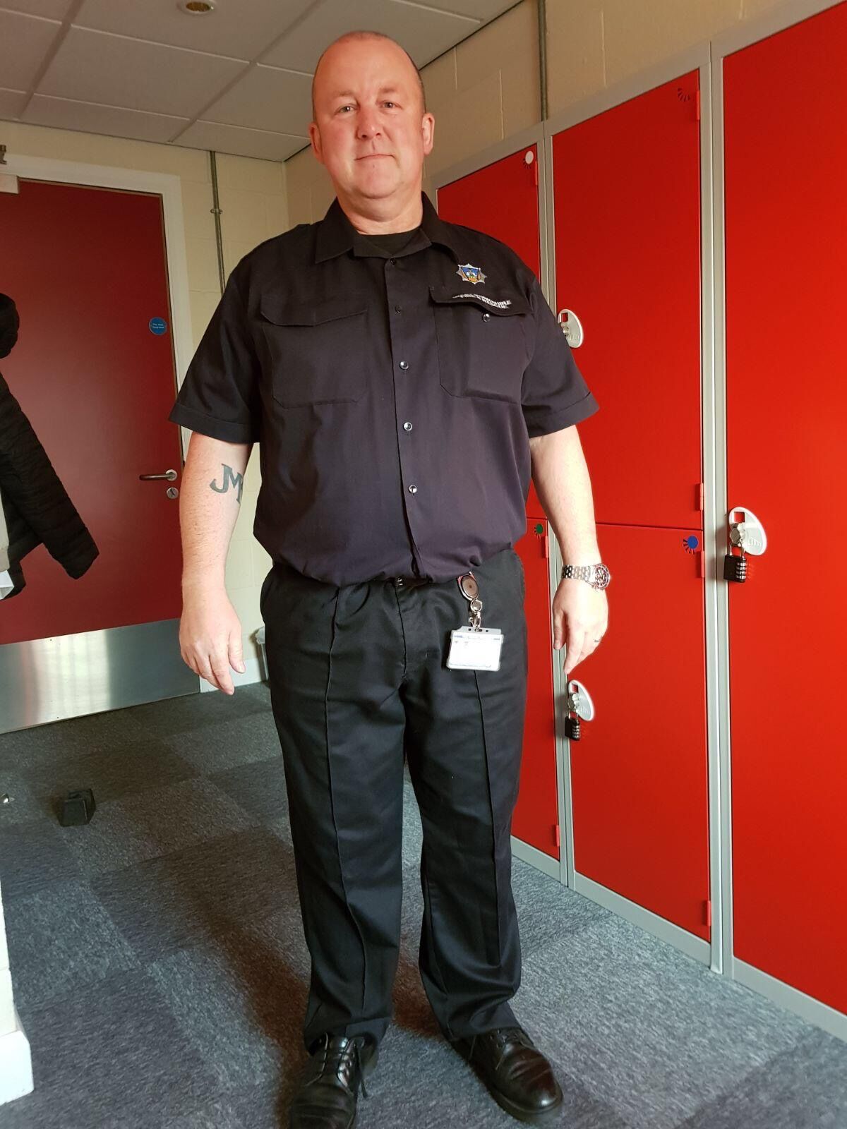 A man in a black uniform stands indoors on a carpeted floor, next to red lockers. He has a visible tattoo on his left forearm and wears a lanyard with an ID badge.