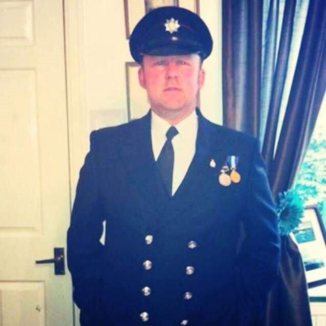A person in a formal dark uniform and peaked cap stands indoors, facing the camera. The uniform has shiny buttons and medals pinned on the chest. There is a closed door and a window with curtains in the background.