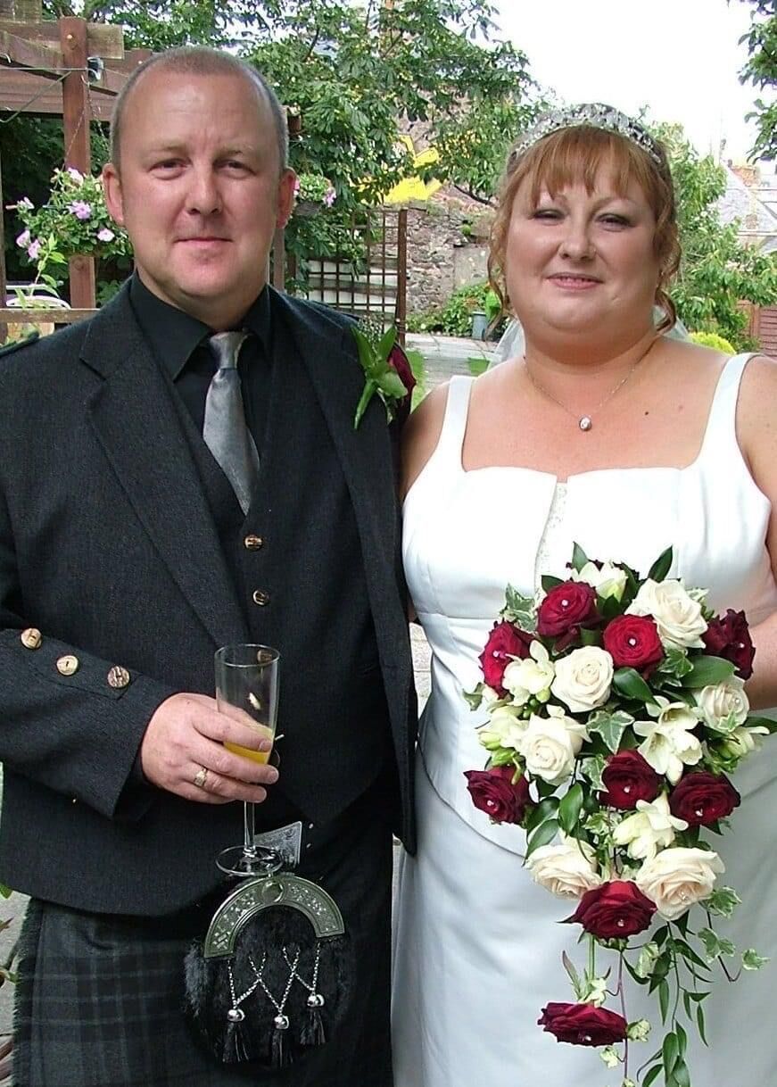 A smiling couple poses outdoors at a wedding. The man wears a dark suit with a kilt and holds a drink. The woman wears a white dress and tiara, holding a bouquet of red and white roses.
