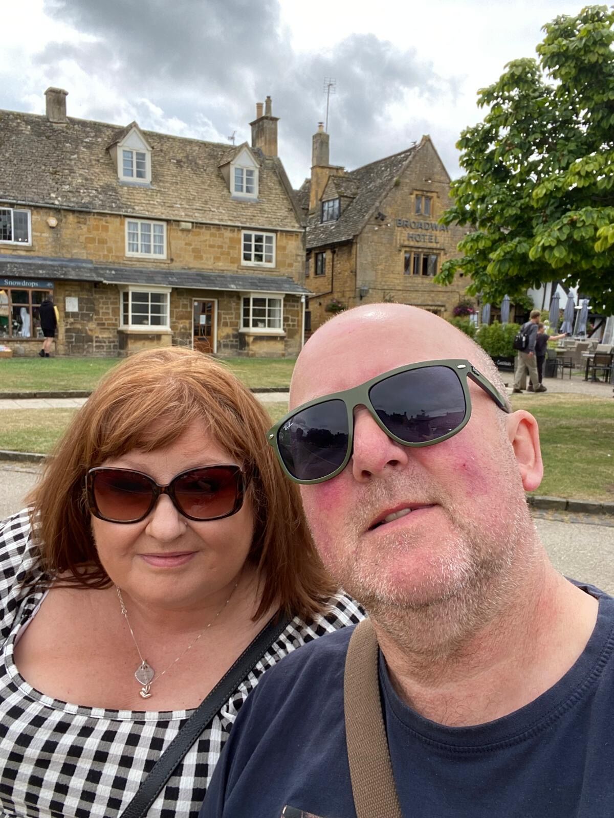 A woman and a man wearing sunglasses take a selfie outdoors in front of historic stone buildings and a hotel sign, with trees and cloudy skies in the background.