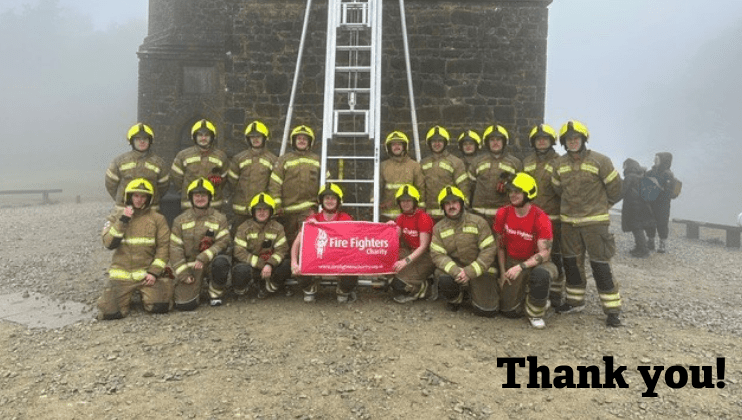 A group of firefighters in uniform and yellow helmets pose outdoors with a red Fire Fighters Charity banner in front of a stone building and tall ladder. 