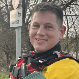 A person wearing a yellow and red rescue suit stands outdoors near a river, smiling at the camera. Behind them are bare trees, a parking area, and a sign that reads 