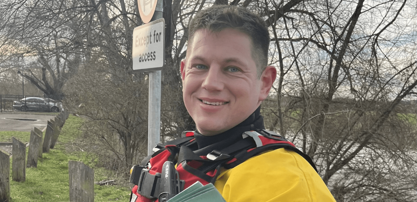 A person wearing a yellow and red rescue suit stands outdoors near a river, smiling at the camera. Behind them are bare trees, a parking area, and a sign that reads 