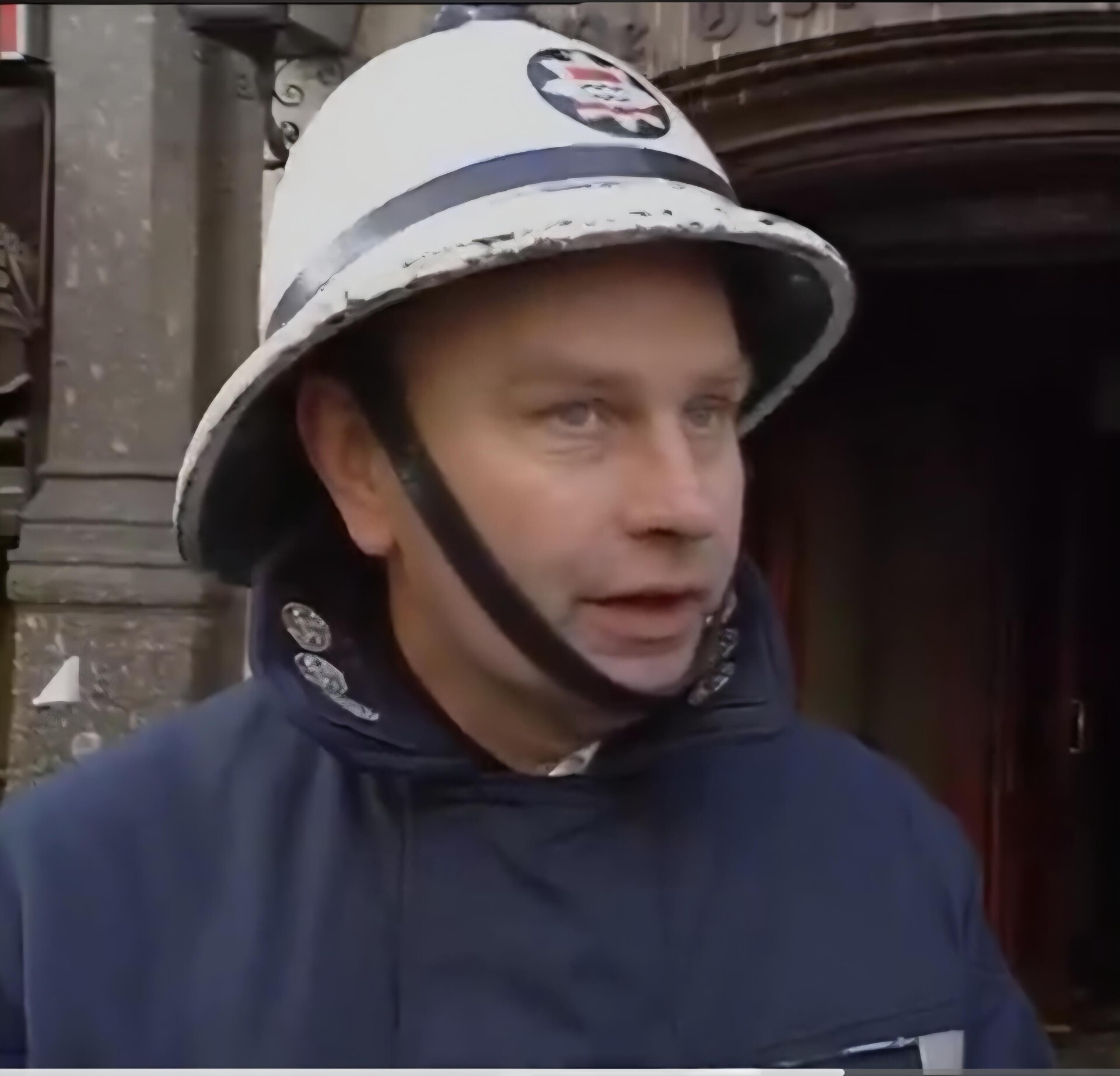 A man wearing a white helmet and a dark uniform stands outside in front of a building, possibly a firefighter or emergency worker, looking slightly to the side.