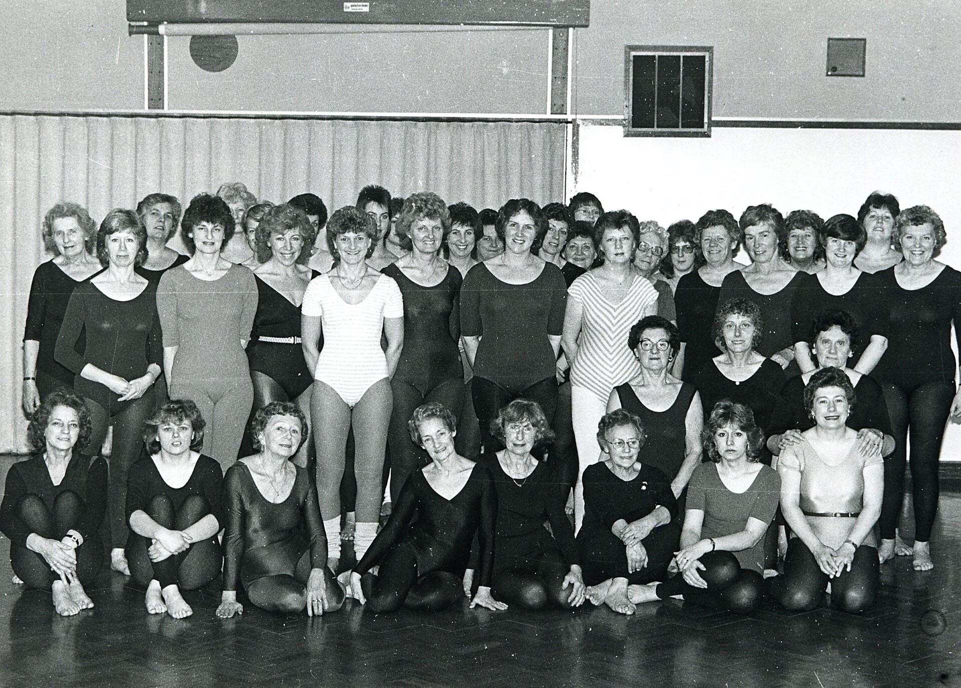 A black-and-white photo of around 30 women in leotards and workout attire posing together in a gymnasium, some standing and some sitting cross-legged on a wooden floor.