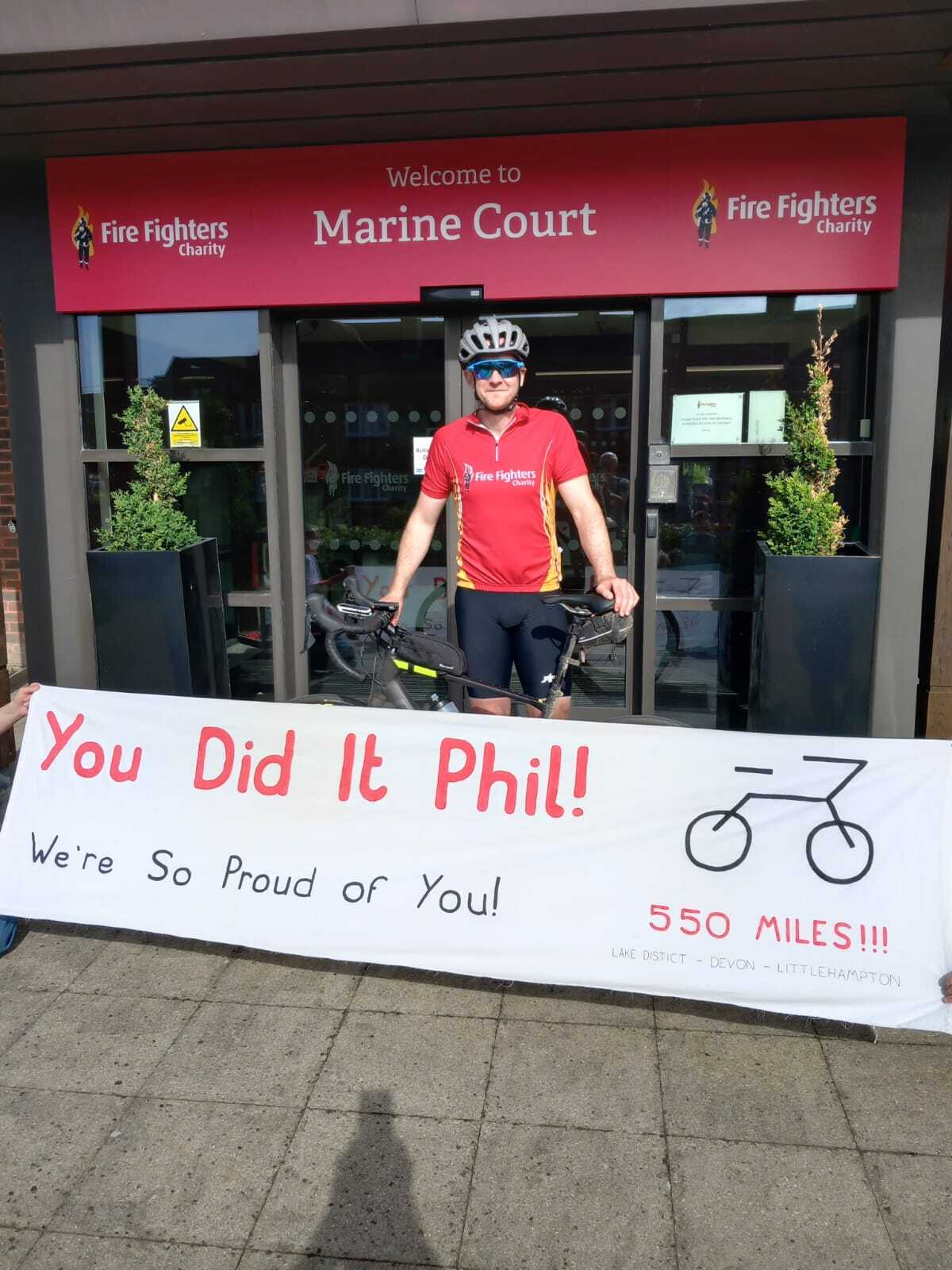 A man in cycling gear stands with his bike outside Marine Court, under a Fire Fighters Charity sign. He smiles behind a large banner that reads, “You Did It Phil! We’re So Proud of You! 550 miles!!!” with a bike illustration.