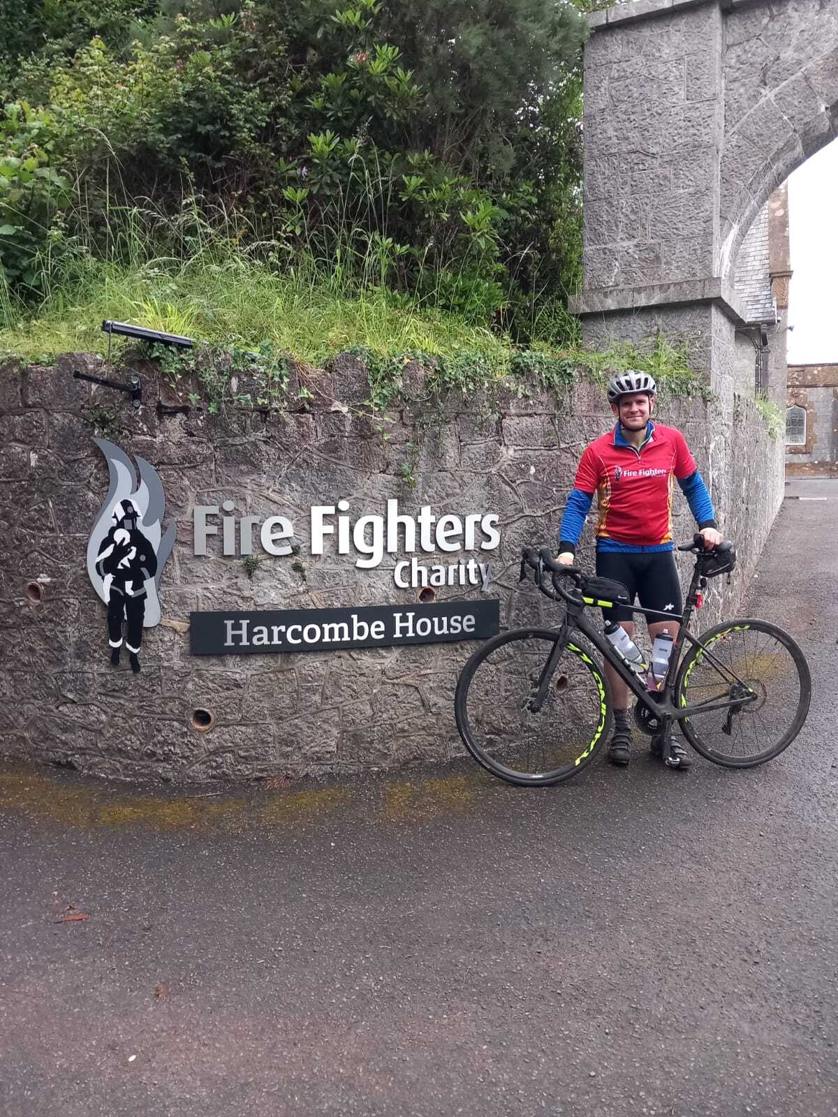 A cyclist in a red jersey and helmet stands with his bike in front of a stone wall with a "Fire Fighters Charity Harcombe House" sign. Trees and an arched stone entrance are visible in the background.