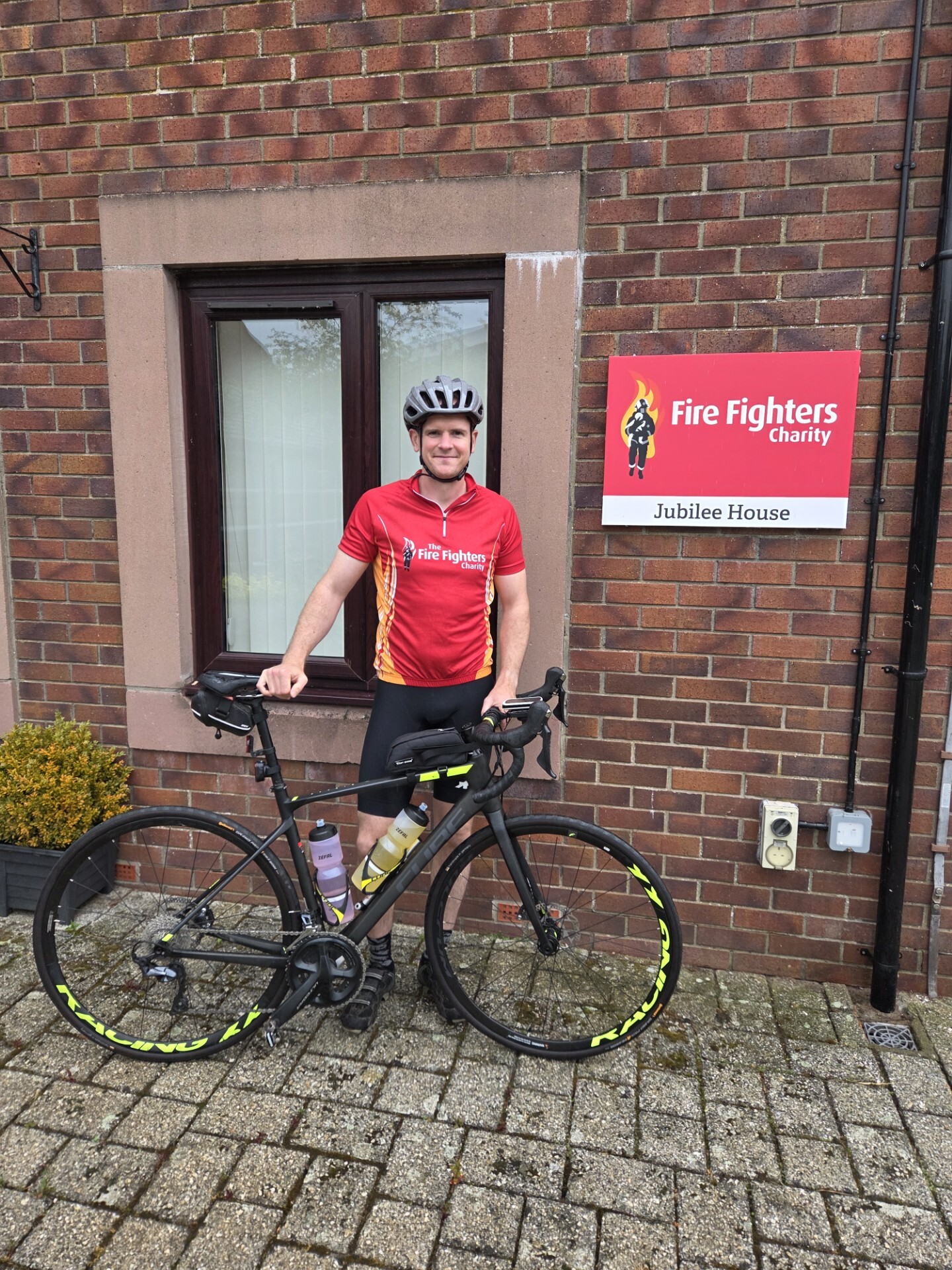 A cyclist in a red "Fire Fighters Charity" jersey and helmet stands with a black road bike outside a brick building, next to a sign reading "Fire Fighters Charity, Jubilee House.
