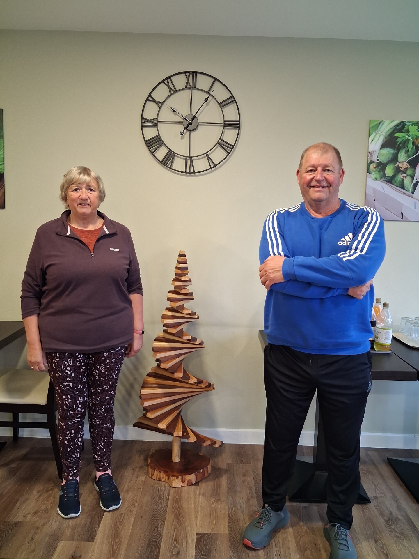 A woman and a man stand indoors on either side of a wooden spiral sculpture shaped like a tree. A large wall clock and two framed photos hang on the beige wall behind them. The room has wooden flooring.