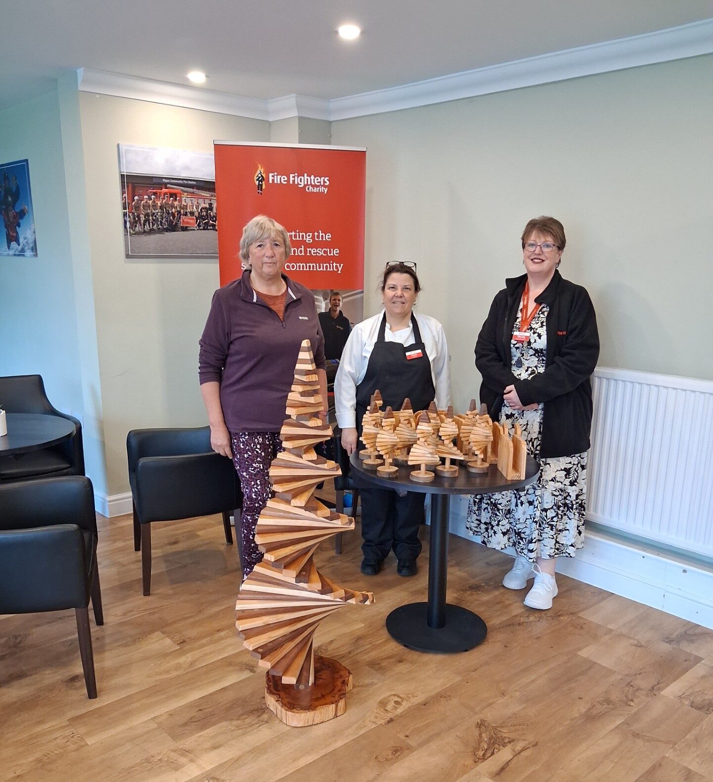 Three women stand indoors beside a table displaying wooden spiral sculptures and small wooden items. A red "Fire Fighters" banner is in the background. The room has wooden floors, chairs, and cream-colored walls.
