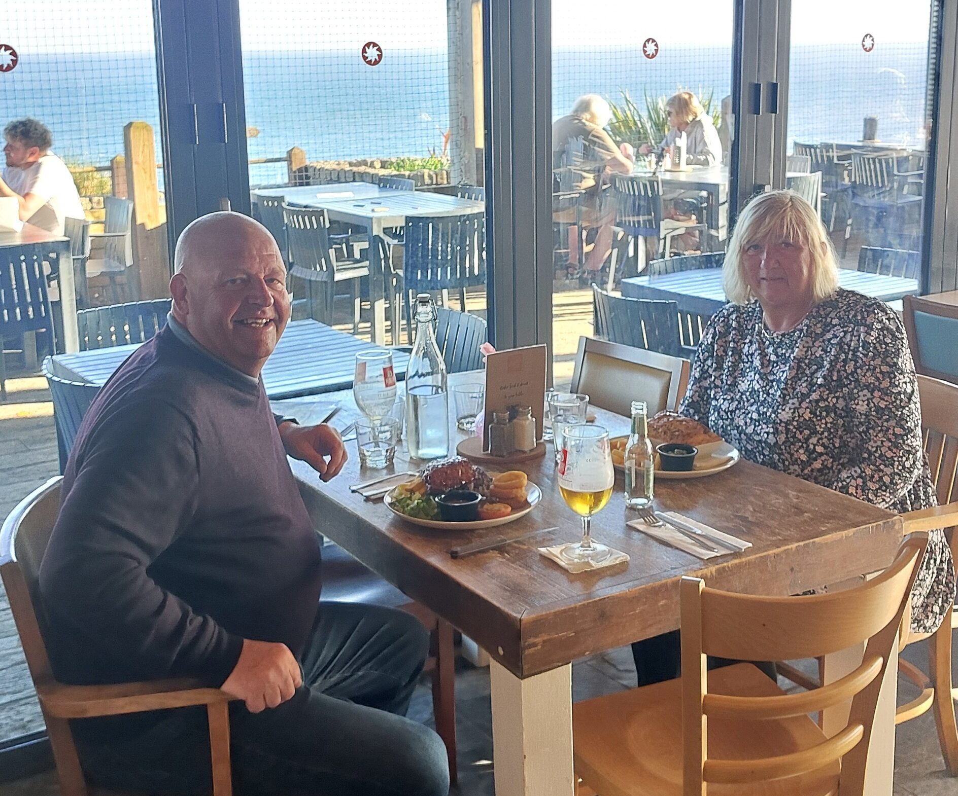 A man and woman sit at a restaurant table by large windows with a seaside view, enjoying a meal and drinks. Sunlight shines through, and other diners are visible on the outdoor terrace.
