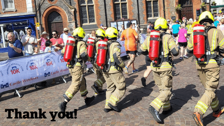 Four firefighters in full gear and helmets run in a public event, with a crowd of spectators and other runners in the background. Banner and buildings are visible. Text on the image reads, 