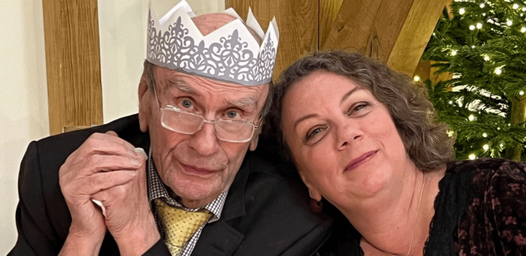 An elderly man wearing glasses, a suit, and a paper crown sits next to a smiling woman with curly hair. They are close together in front of a decorated Christmas tree.