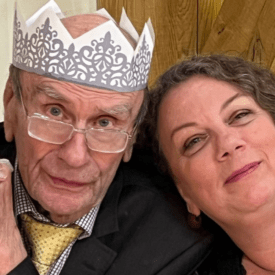An elderly man wearing glasses, a suit, and a paper crown sits next to a smiling woman with curly hair. They are close together in front of a decorated Christmas tree.
