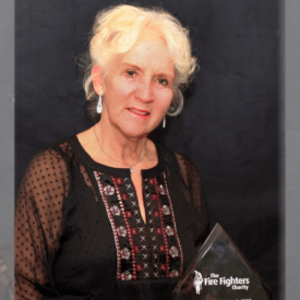 An older woman with curly white hair, wearing a black embroidered blouse, stands in front of a dark backdrop holding a glass award that reads 