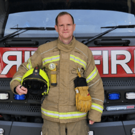 A firefighter in full uniform stands holding a helmet and gloves in front of a red fire truck, with the word 