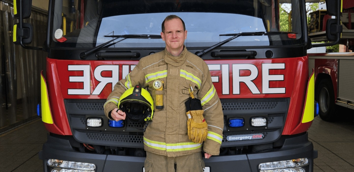 A firefighter in full uniform stands holding a helmet and gloves in front of a red fire truck, with the word 