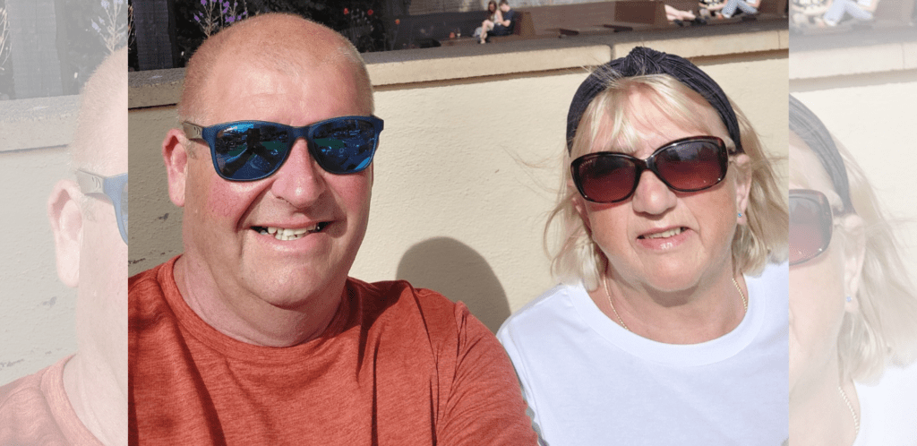 A smiling man in a red shirt and a woman in a white shirt, both wearing sunglasses, sit outside in bright sunlight with a beige wall behind them.