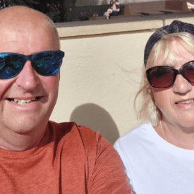 A smiling man in a red shirt and a woman in a white shirt, both wearing sunglasses, sit outside in bright sunlight with a beige wall behind them.