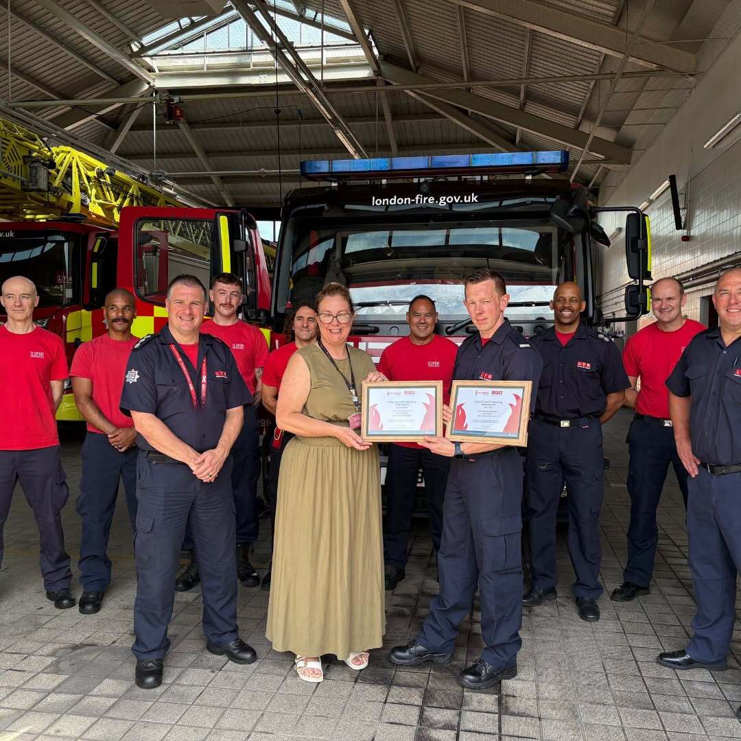A group of firefighters in uniform stand smiling in a fire station with fire trucks behind them. A woman in a beige dress and a firefighter hold certificates in the center of the group.