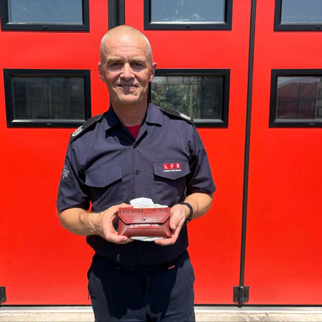 A smiling firefighter in uniform stands in front of bright red doors, holding a small wrapped gift with tissue paper visible.