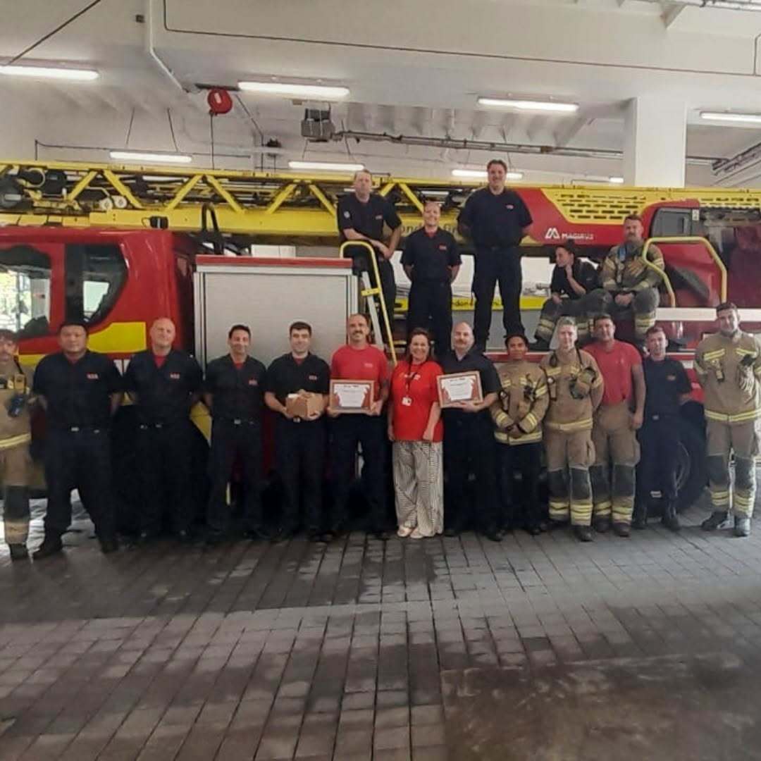 A group of firefighters in uniform pose in front of a large red fire truck inside a fire station. Two people in the center hold certificates, and everyone is smiling for the photo.